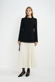 Angled view of a model in an ecru pleated midi skirt beneath a buttoned black collarless jacket, paired with dark socks and pointed black shoes.