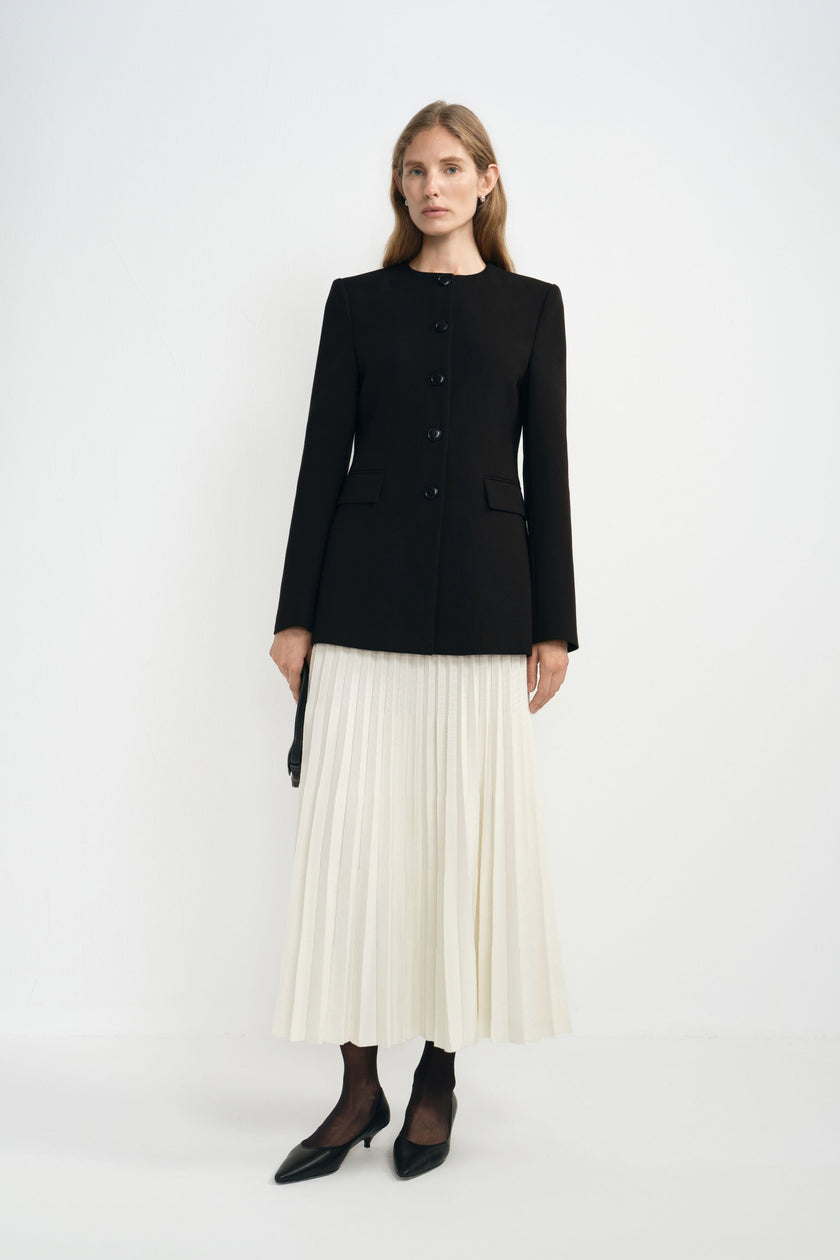 Angled view of a model in an ecru pleated midi skirt beneath a buttoned black collarless jacket, paired with dark socks and pointed black shoes.