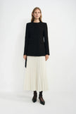 A model wearing an ecru wool-blend midi skirt with narrow accordion pleats, paired with a black collarless jacket, dark socks and pointed black shoes.