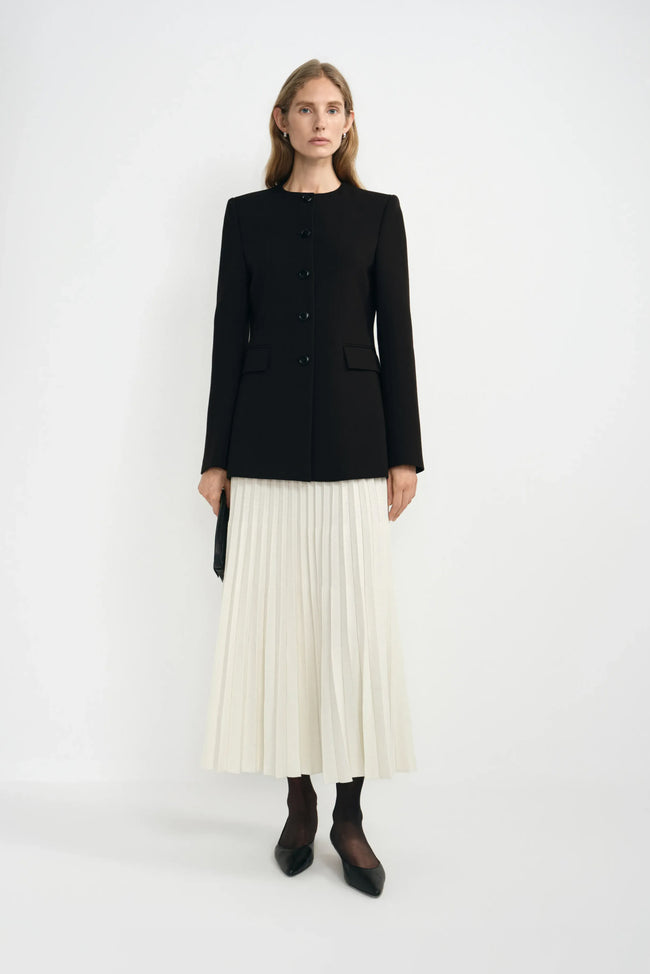 A model wearing an ecru wool-blend midi skirt with narrow accordion pleats, paired with a black collarless jacket, dark socks and pointed black shoes.