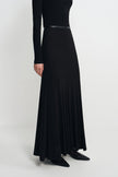 A model wearing a long black maxi dress with a thin black belt at the waist, styled with black pointed-toe flats against a light gray studio background.