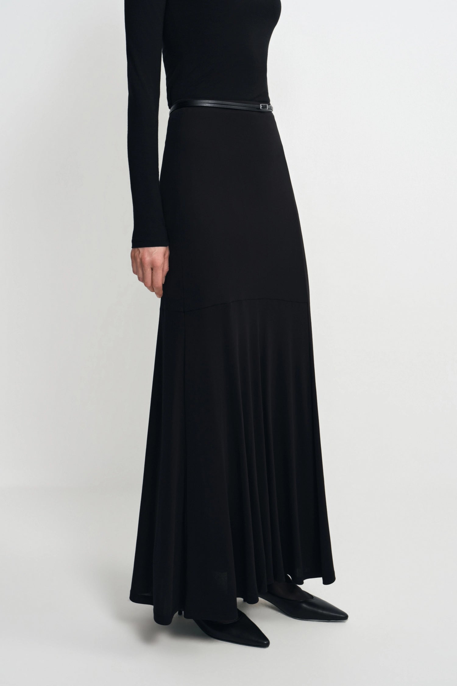 A model wearing a long black maxi dress with a thin black belt at the waist, styled with black pointed-toe flats against a light gray studio background.