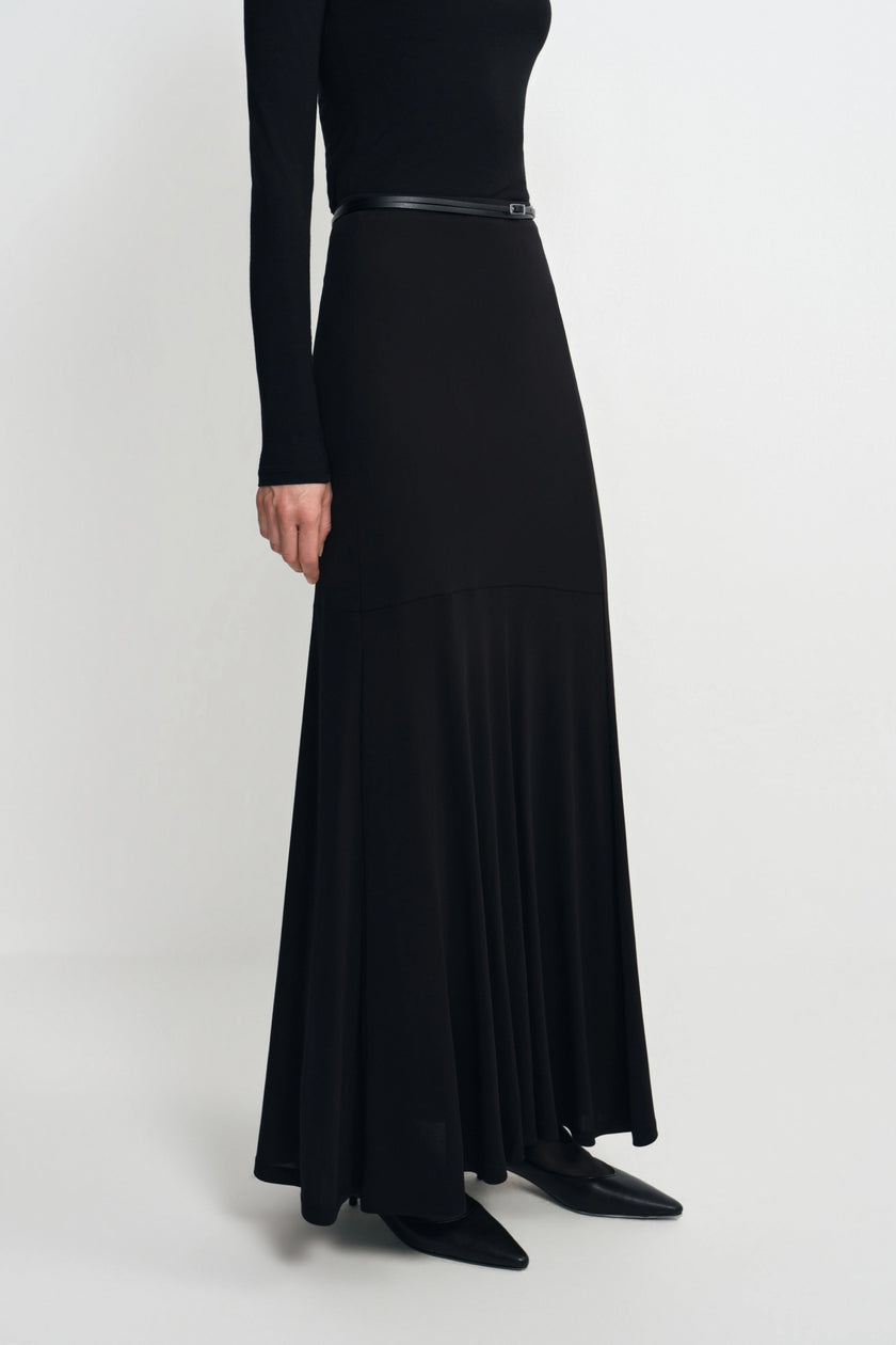 A model wearing a long black maxi dress with a thin black belt at the waist, styled with black pointed-toe flats against a light gray studio background.