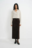 A model stands against a plain white studio background wearing a cream and beige plaid button-up shirt, a dark brown midi skirt, a thin black belt, sheer black ankle socks, and black Mary Jane shoes.