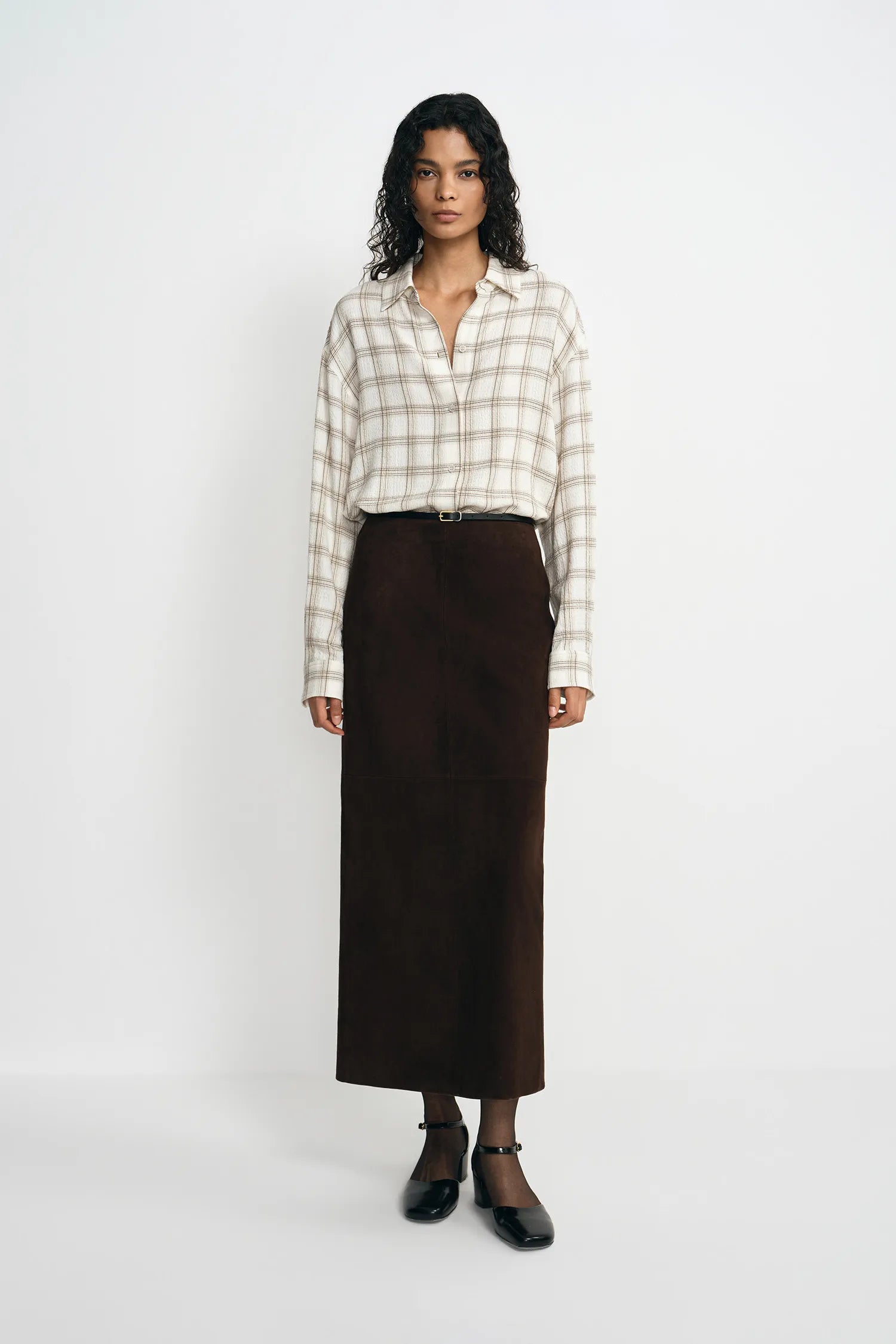A model stands against a plain white studio background wearing a cream and beige plaid button-up shirt, a dark brown midi skirt, a thin black belt, sheer black ankle socks, and black Mary Jane shoes.