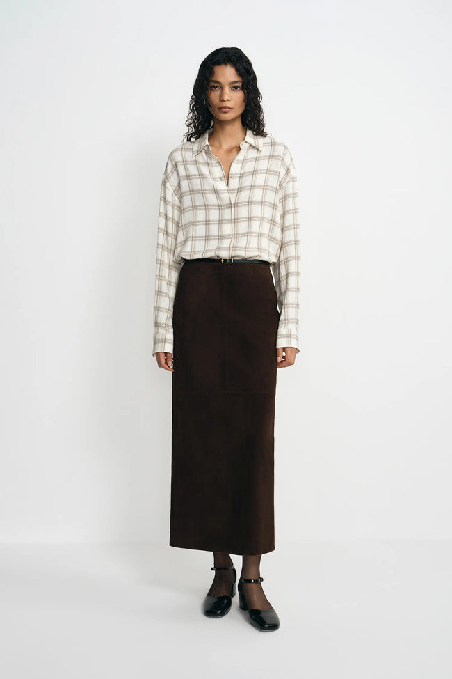 A model stands against a plain white studio background wearing a cream and beige plaid button-up shirt, a dark brown midi skirt, a thin black belt, sheer black ankle socks, and black Mary Jane shoes.