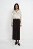 A model stands against a plain white studio background wearing a cream and beige plaid button-up shirt, a dark brown midi skirt, a thin black belt, sheer black ankle socks, and black Mary Jane shoes.