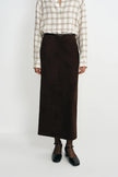 A model is shown from the neck down wearing a dark brown midi skirt, a beige and brown plaid button-up shirt, and black Mary Jane shoes against a plain white studio background.