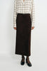 A model is shown from the neck down wearing a dark brown midi skirt, a beige and brown plaid button-up shirt, and black Mary Jane shoes against a plain white studio background.