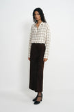 A model stands against a light gray studio background wearing a cream and beige plaid button-up shirt, a dark brown wide-leg skirt, and black Mary Jane heels.