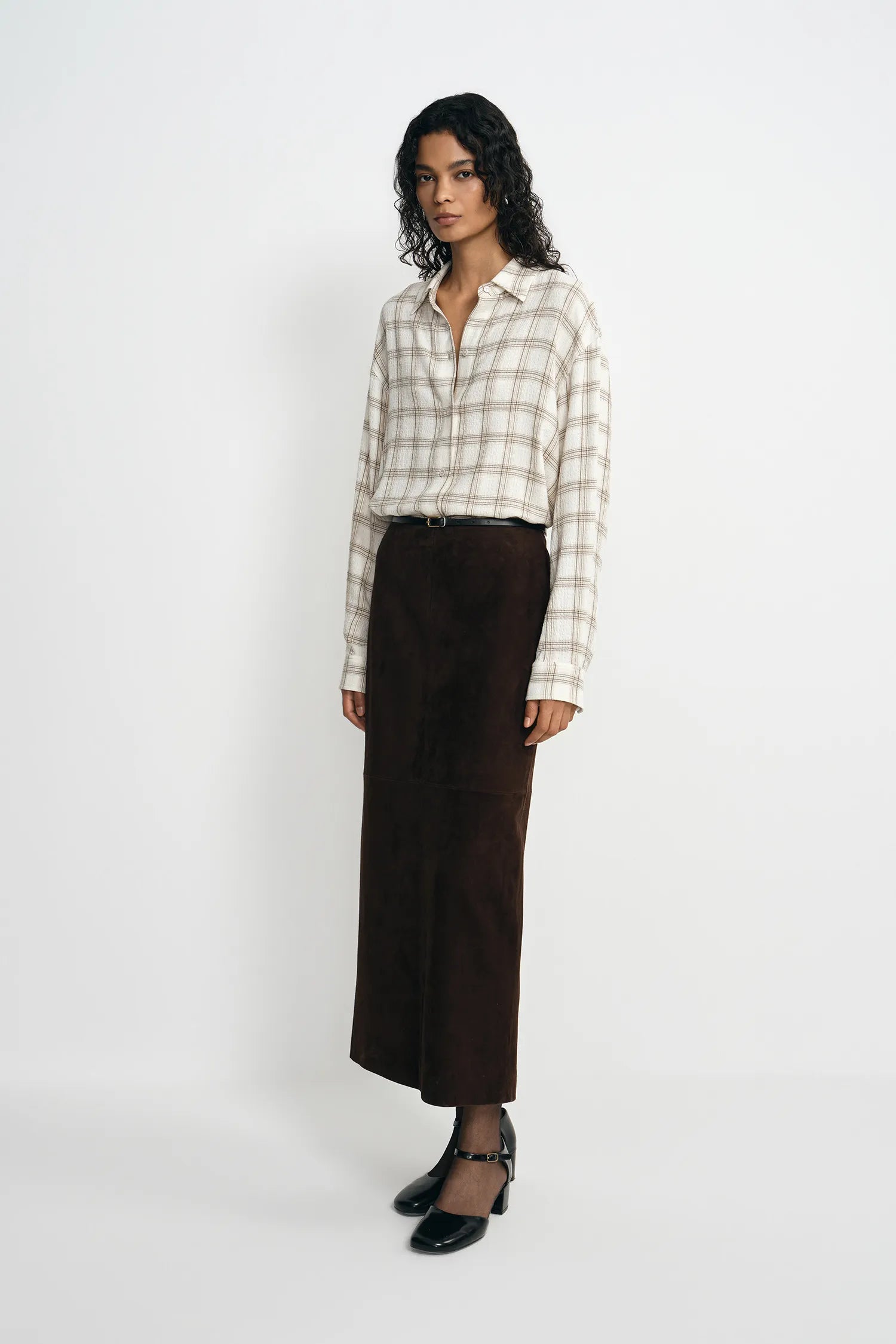 A model stands against a light gray studio background wearing a cream and beige plaid button-up shirt, a dark brown wide-leg skirt, and black Mary Jane heels.