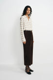 A model stands against a plain light gray studio background wearing a cream and beige plaid long-sleeve button-up shirt, a dark brown midi skirt, sheer black tights, and black pointed flats.