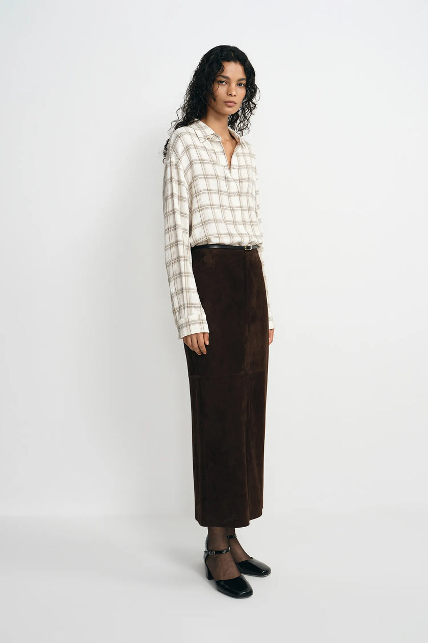 A model stands against a plain light gray studio background wearing a cream and beige plaid long-sleeve button-up shirt, a dark brown midi skirt, sheer black tights, and black pointed flats.