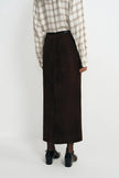 A person shown from the back wearing a dark brown midi skirt, a beige and brown plaid shirt, and black shoes against a plain light gray studio background.