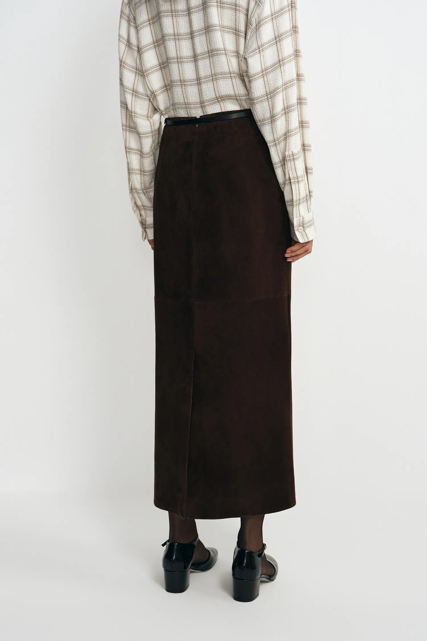 A person shown from the back wearing a dark brown midi skirt, a beige and brown plaid shirt, and black shoes against a plain light gray studio background.