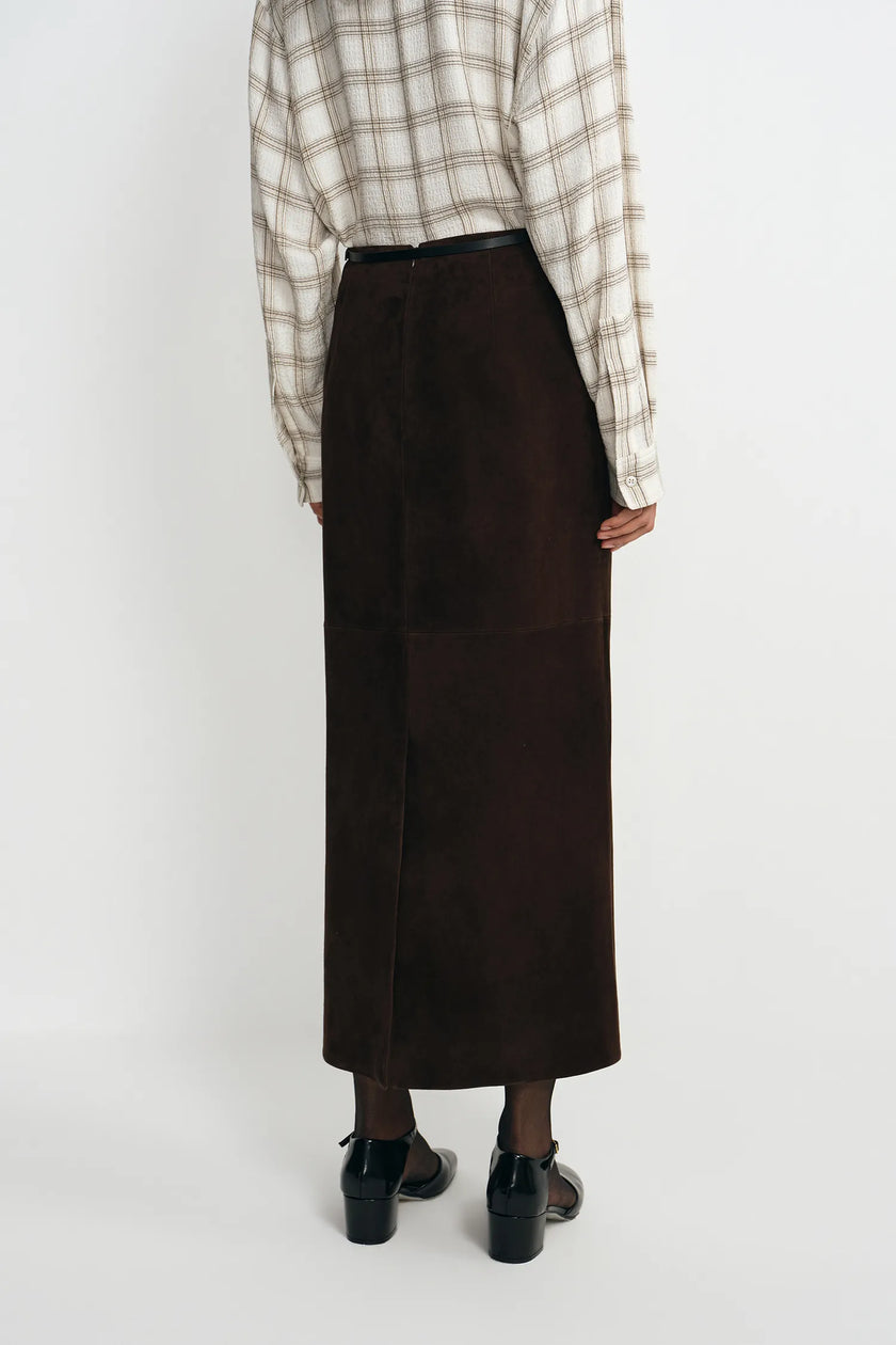 A person shown from the back wearing a dark brown midi skirt, a beige and brown plaid shirt, and black shoes against a plain light gray studio background.