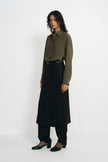 Three-quarter view of a model in a black layered-panel midi skirt worn over black trousers, paired with an olive shirt tucked into a belted waist.