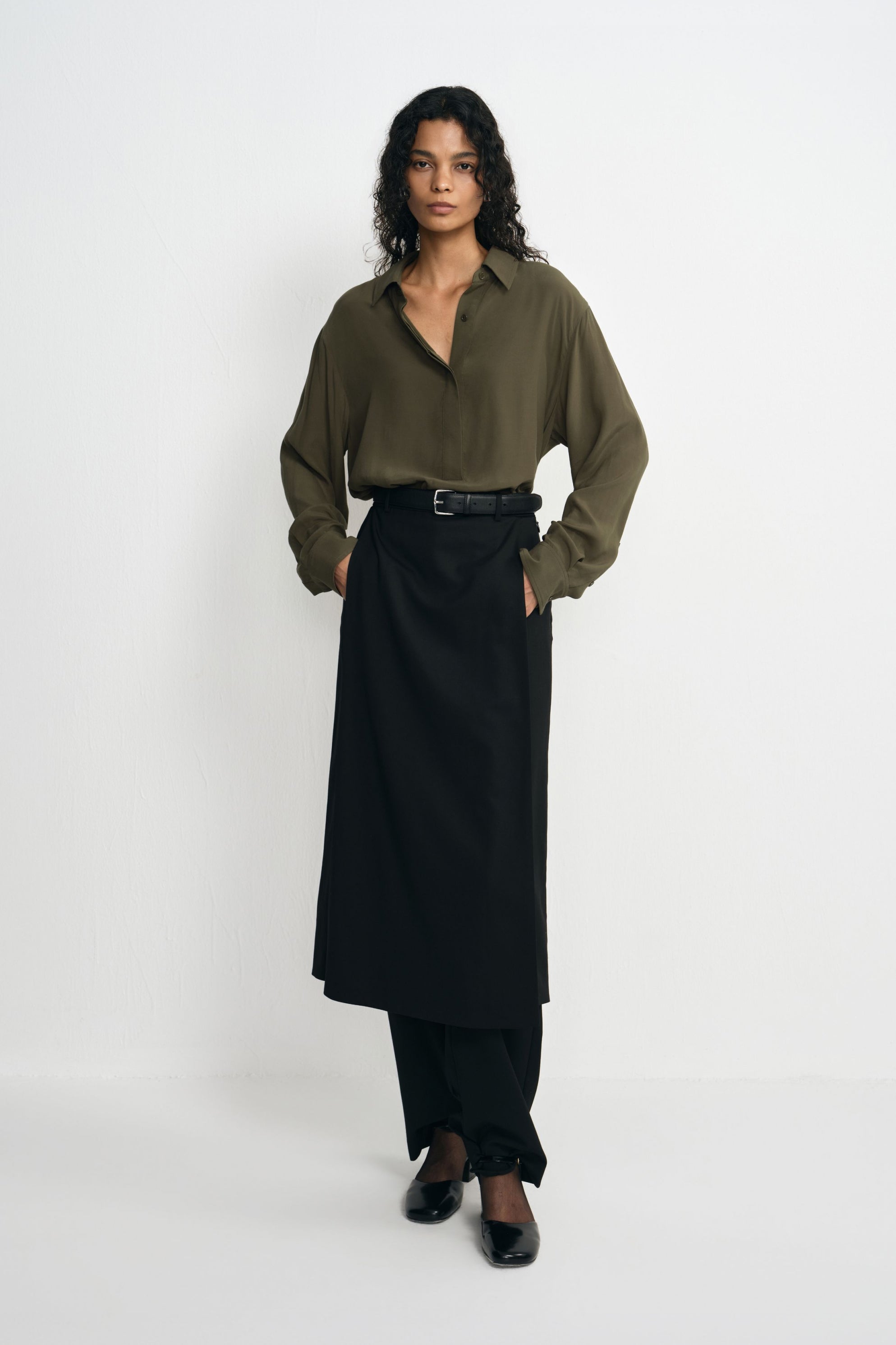 A model wearing a black wool-blend midi skirt over trousers, with hands in the pockets showing the front panel's drape beneath a tucked olive shirt.