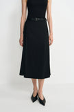 Close-up of a black midi skirt on a model, showing the overlapping front panel and calf-length hem, styled with a black tank, belt and pointed shoes.