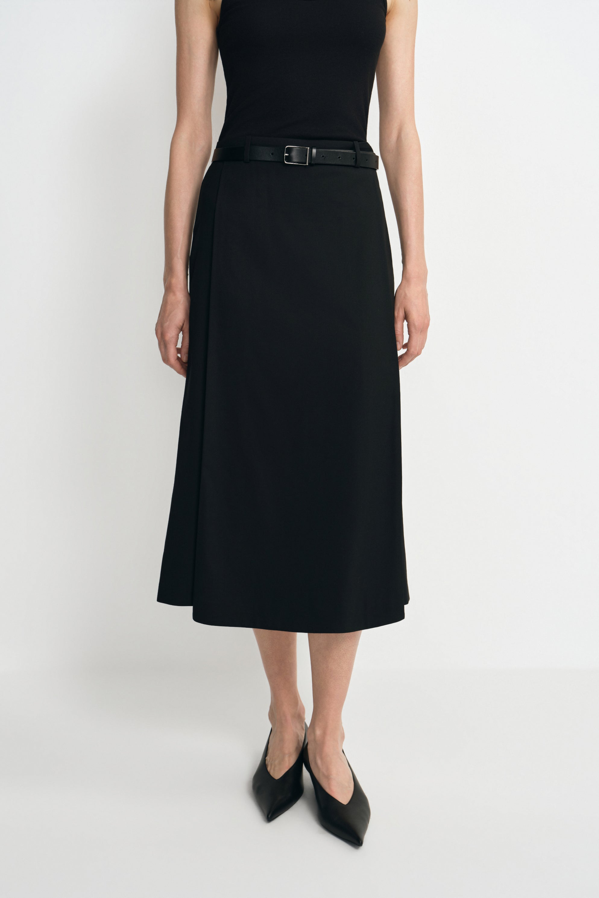 Close-up of a black midi skirt on a model, showing the overlapping front panel and calf-length hem, styled with a black tank, belt and pointed shoes.
