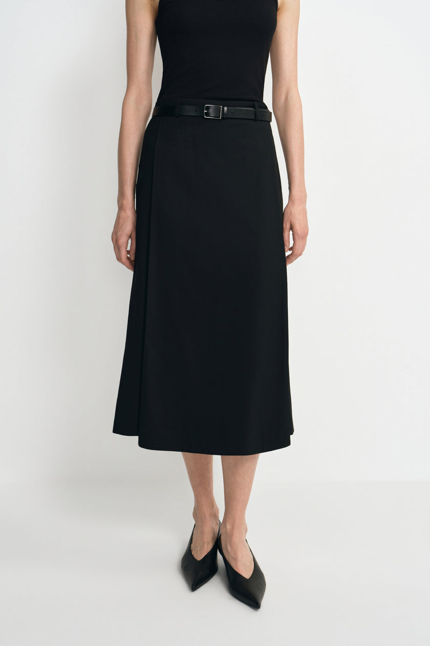 Close-up of a black midi skirt on a model, showing the overlapping front panel and calf-length hem, styled with a black tank, belt and pointed shoes.
