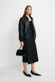 Angled view of a model in a black layered-panel midi skirt, paired with an open black bomber jacket, pointed shoes and a structured black handbag.