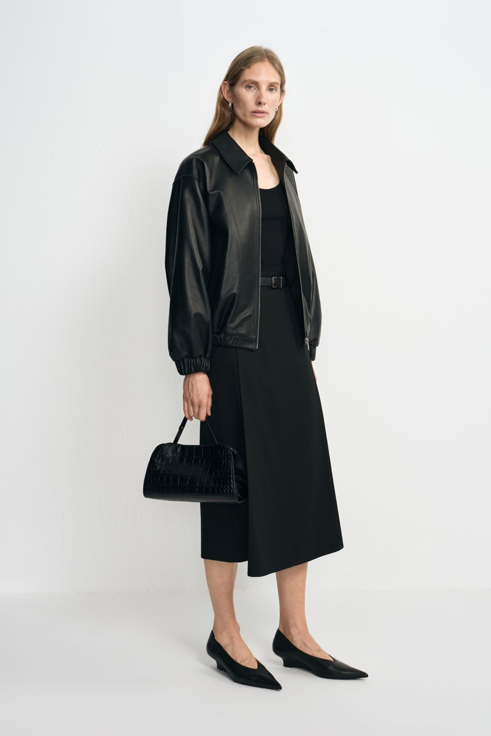 Angled view of a model in a black layered-panel midi skirt, paired with an open black bomber jacket, pointed shoes and a structured black handbag.
