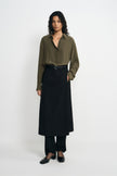 A model facing forward in a black panelled midi skirt layered over trousers, paired with a tucked olive shirt, black belt and square-toe shoes.