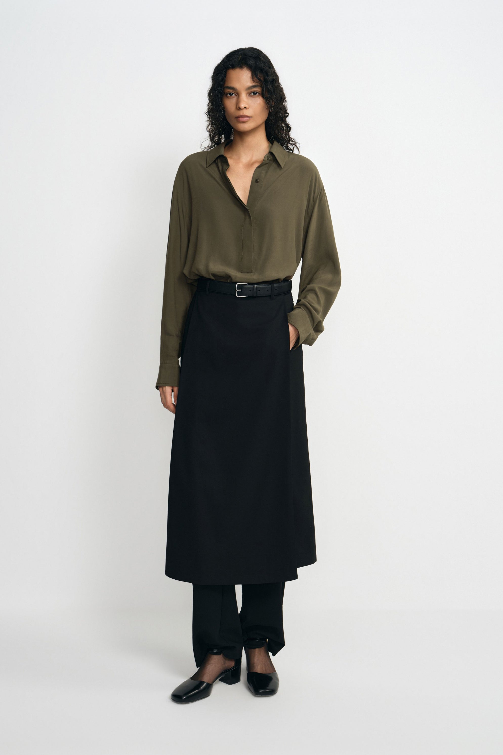 A model facing forward in a black panelled midi skirt layered over trousers, paired with a tucked olive shirt, black belt and square-toe shoes.