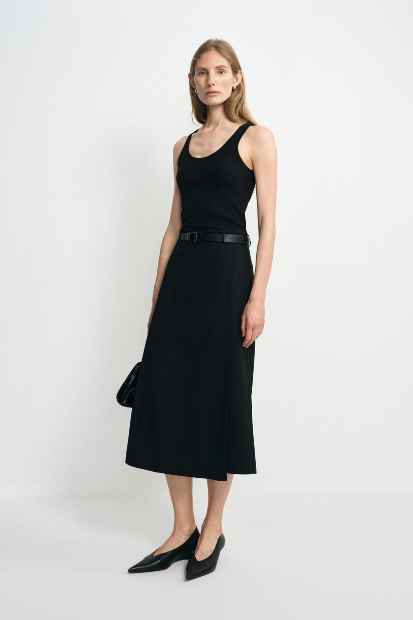Three-quarter view of a model wearing a black wool-blend midi skirt with an overlapping panel, styled with a black tank, slim belt and pointed shoes.