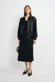 A model wearing a black layered-panel midi skirt with a flared hem, paired with a black tank, open bomber jacket and pointed black shoes.