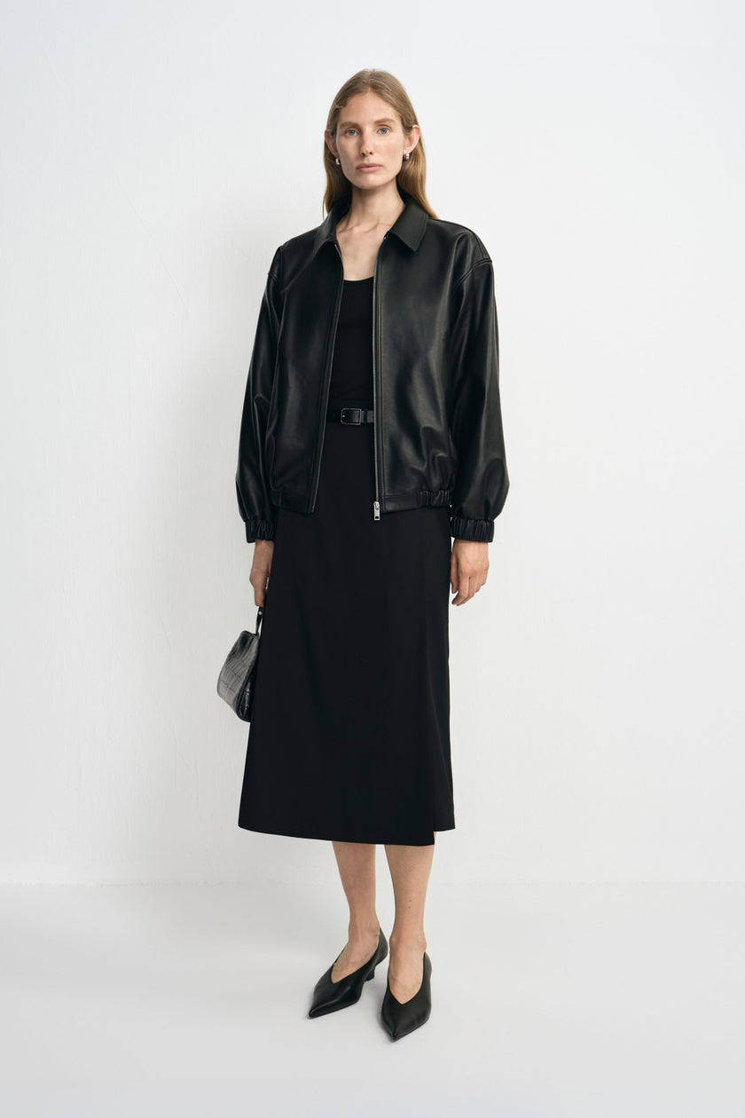 A model wearing a black layered-panel midi skirt with a flared hem, paired with a black tank, open bomber jacket and pointed black shoes.