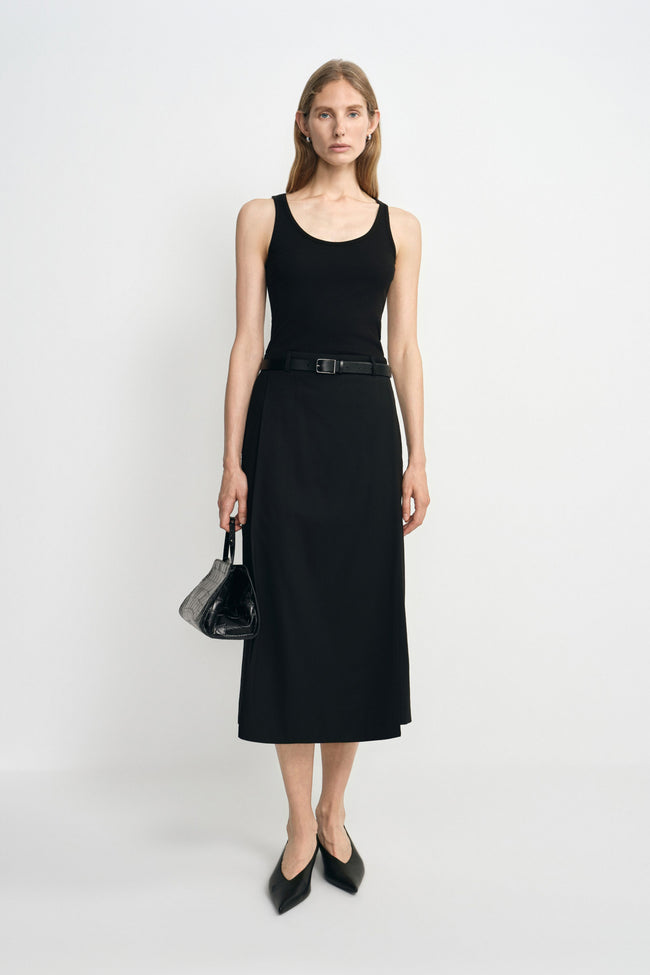 A model wearing a black wool-blend midi skirt with an overlapping front panel, styled with a black tank, narrow belt, pointed shoes and small handbag.