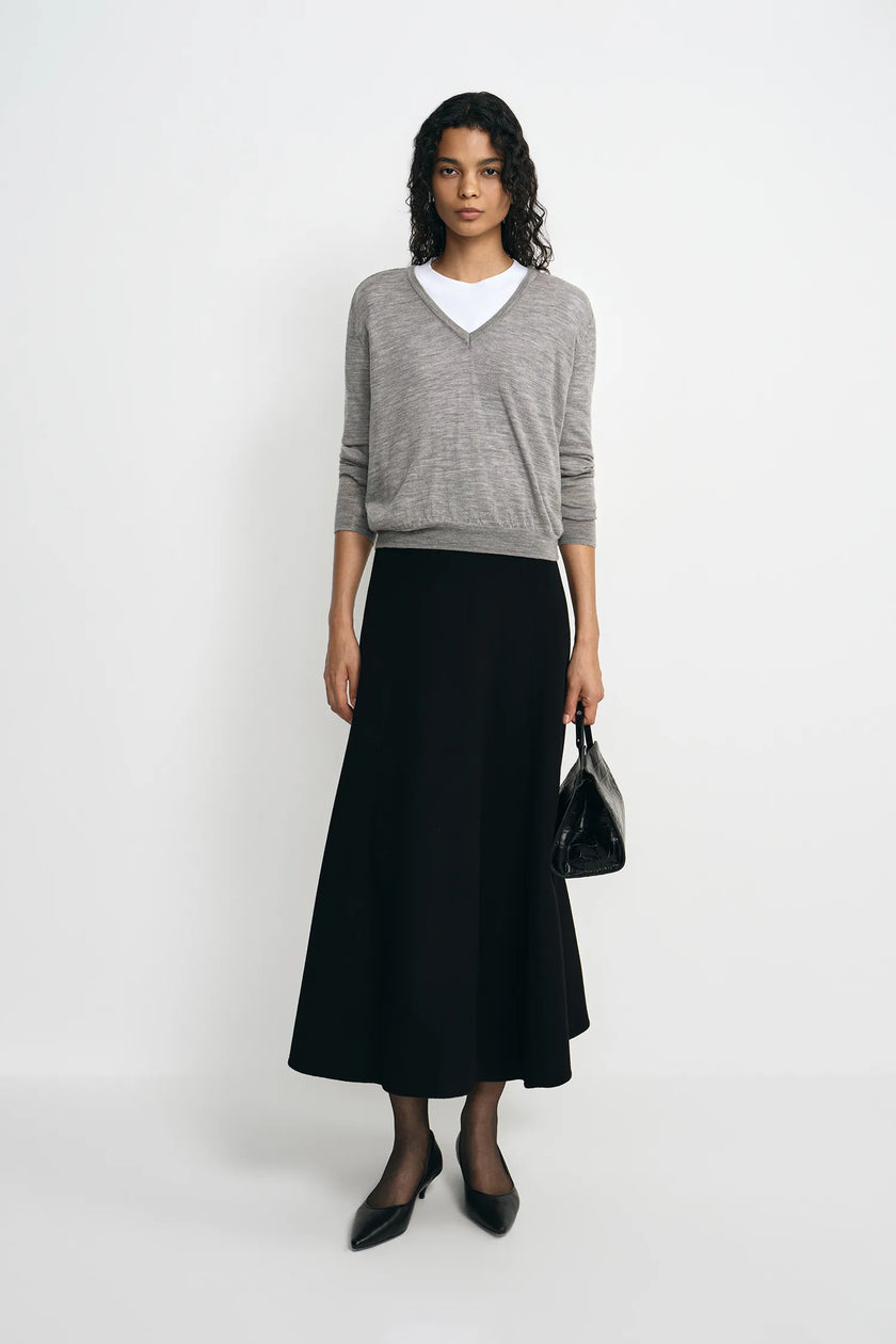 A woman stands against a light gray studio background wearing a heather gray V-neck sweater over a white crew-neck top, a long black skirt, black pointed shoes, and holding a small black handbag.