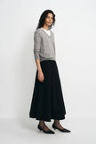 A woman modeling a light gray knit sweater layered over a white crewneck top, paired with a long black skirt and black pointed flats, against a plain white studio background.
