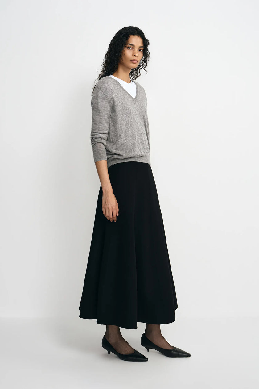 A woman modeling a light gray knit sweater layered over a white crewneck top, paired with a long black skirt and black pointed flats, against a plain white studio background.