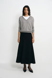 A model stands against a light gray studio background wearing a gray V-neck knit sweater over a white crew-neck undershirt, paired with a long black skirt and black pointed flats.