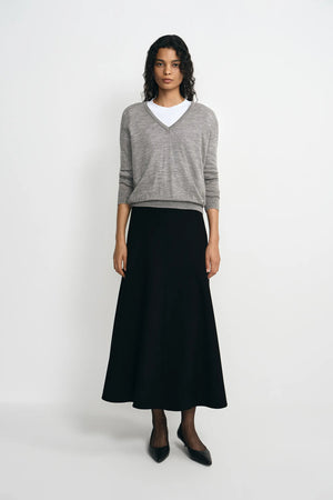 A model stands against a light gray studio background wearing a gray V-neck knit sweater over a white crew-neck undershirt, paired with a long black skirt and black pointed flats.