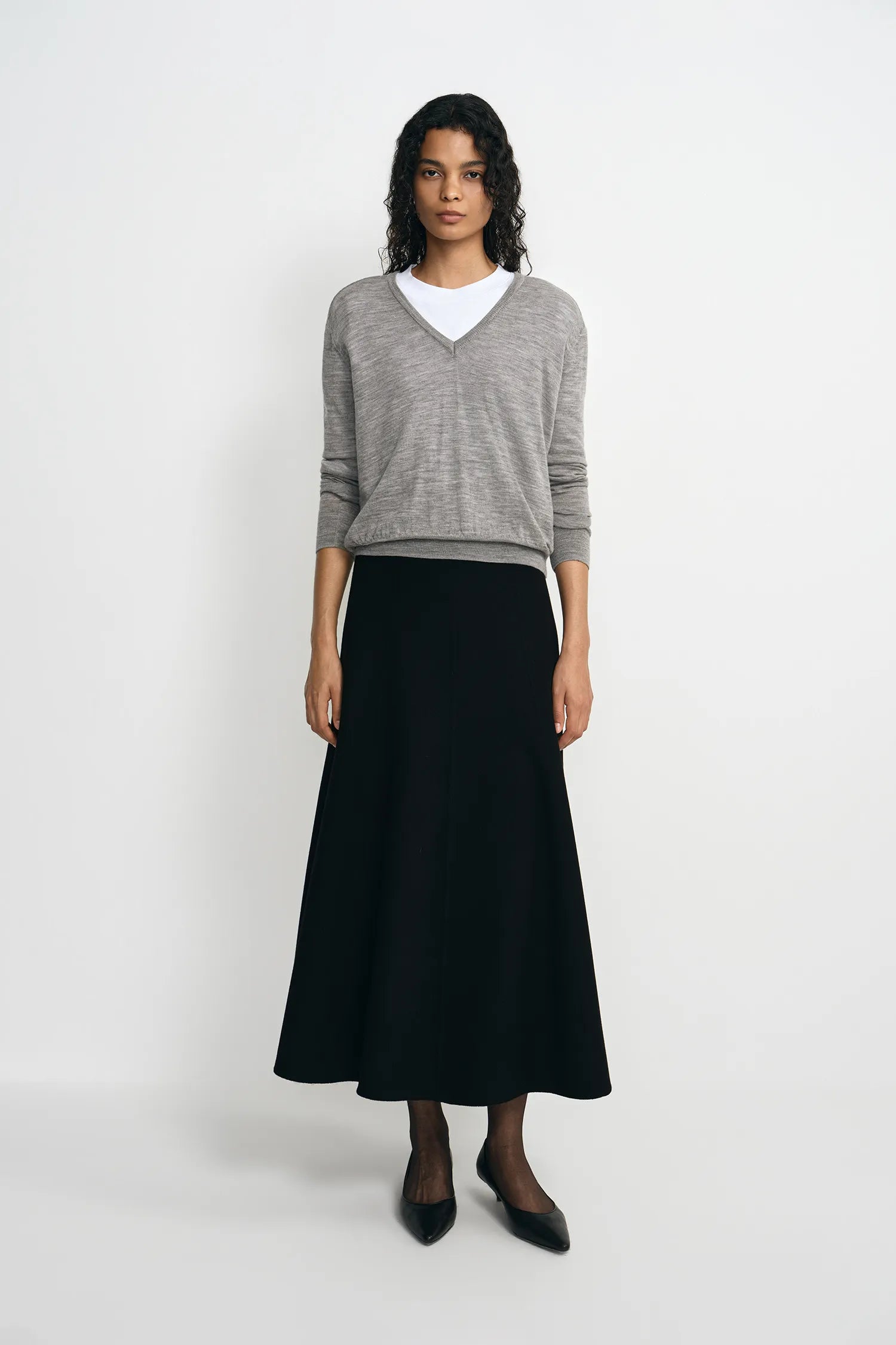 A model stands against a light gray studio background wearing a gray V-neck knit sweater over a white crew-neck undershirt, paired with a long black skirt and black pointed flats.