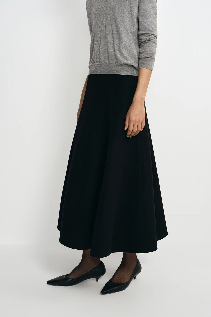 A model wearing a black A-line midi skirt paired with a gray long-sleeve top, sheer black tights, and black pointed-toe kitten heels, shown against a plain light gray studio background.