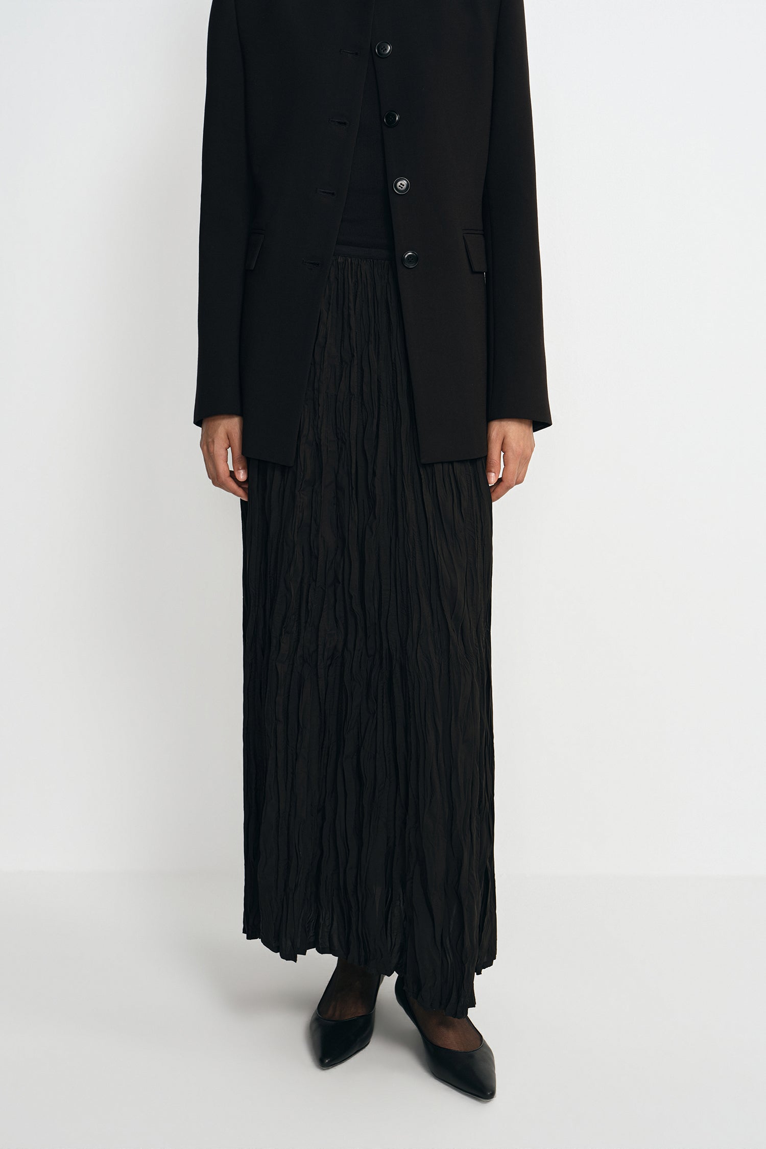 Close-up of a black crinkled maxi skirt showing the elastic waistband, paired with a collarless button-front jacket.