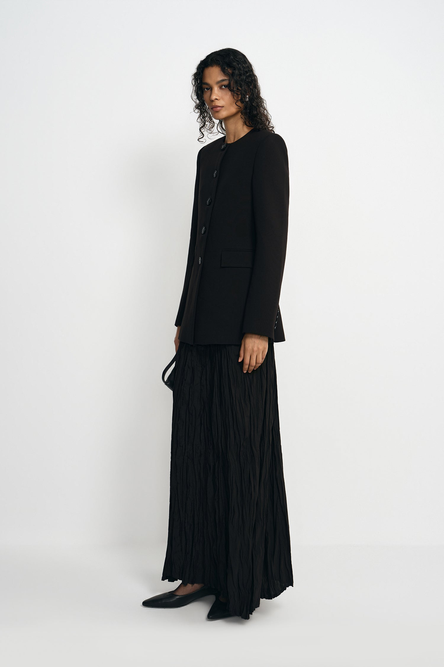 A model wearing a black crinkled maxi skirt, pairing a collarless button-front jacket, side view.
