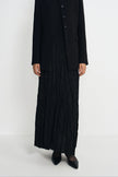A model wearing a black pleated maxi skirt, a black pleated vest, and a black blazer with black pointed-toe heels, photographed against a seamless white studio background.