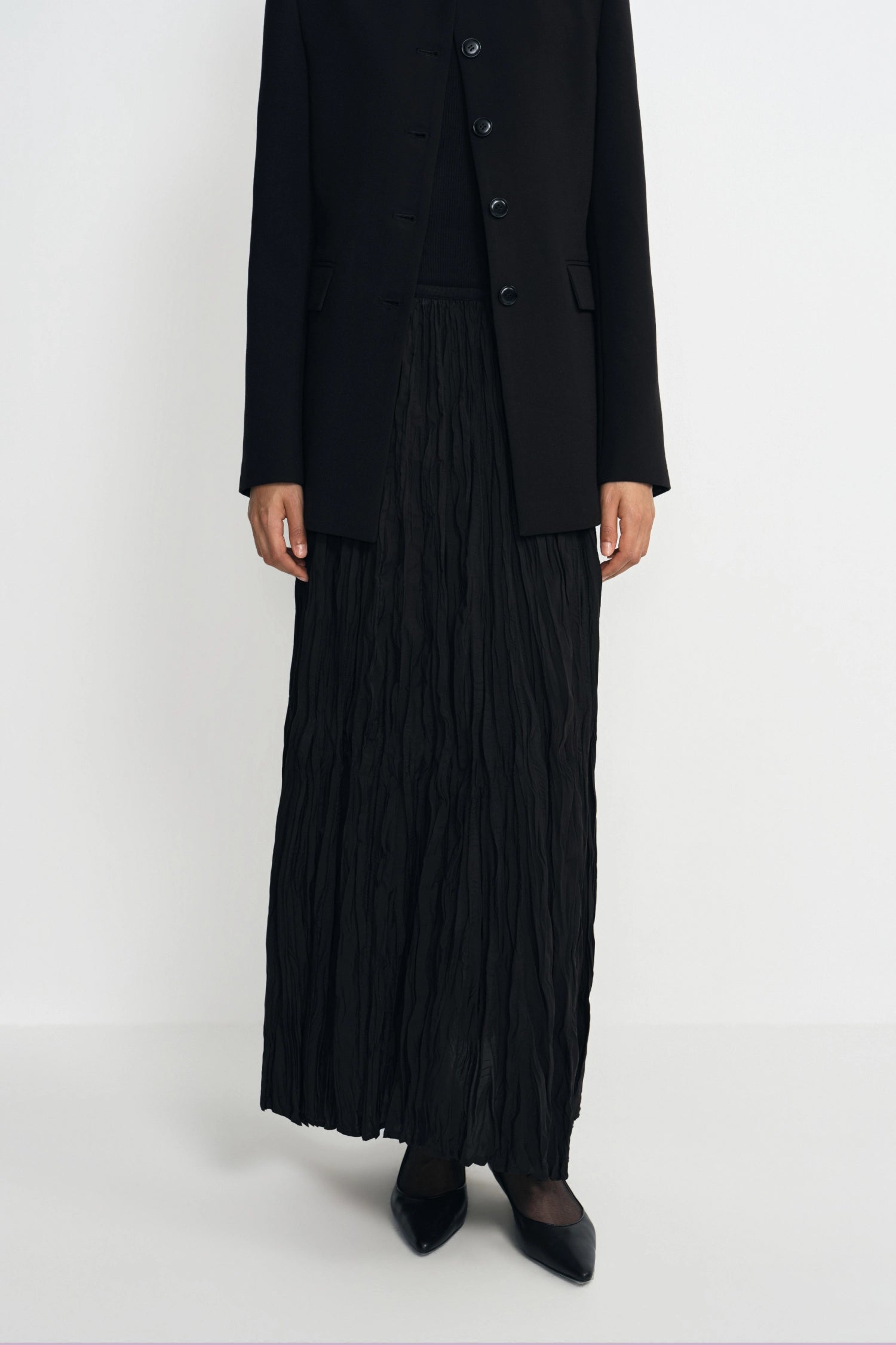 A model wearing a black pleated maxi skirt, a black pleated vest, and a black blazer with black pointed-toe heels, photographed against a seamless white studio background.