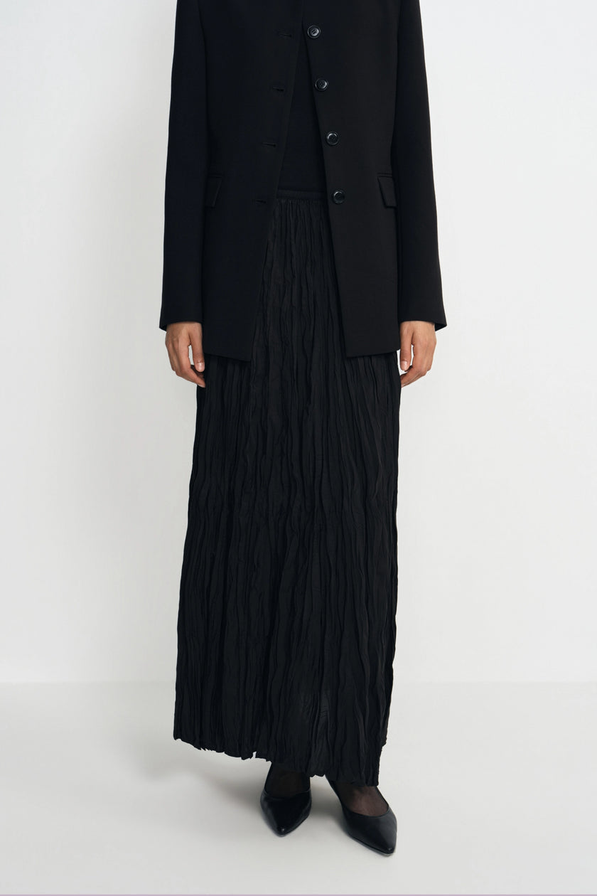 A model wearing a black pleated maxi skirt, a black pleated vest, and a black blazer with black pointed-toe heels, photographed against a seamless white studio background.