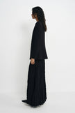 A model shown from a side profile wearing a black long-sleeve cropped blazer and a matching black pleated maxi skirt, standing against a plain white studio background.