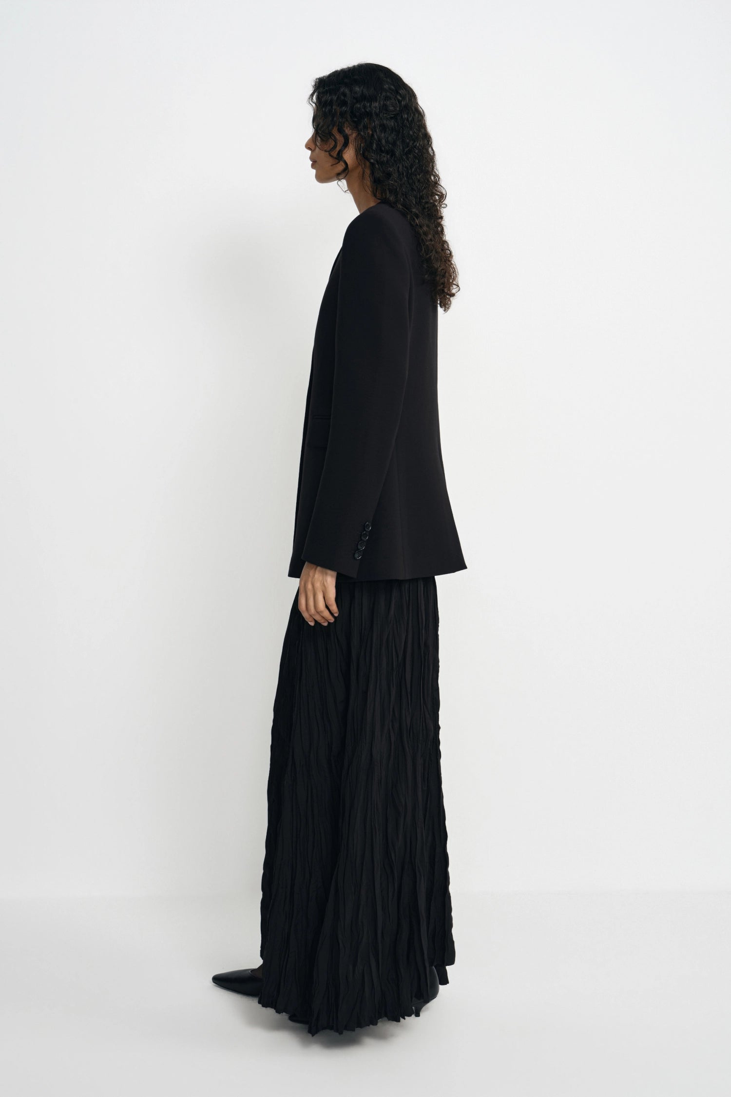 A model shown from a side profile wearing a black long-sleeve cropped blazer and a matching black pleated maxi skirt, standing against a plain white studio background.