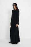 A model stands in a studio wearing a black long-sleeve cropped jacket and a long black pleated skirt, photographed against a plain light gray background.