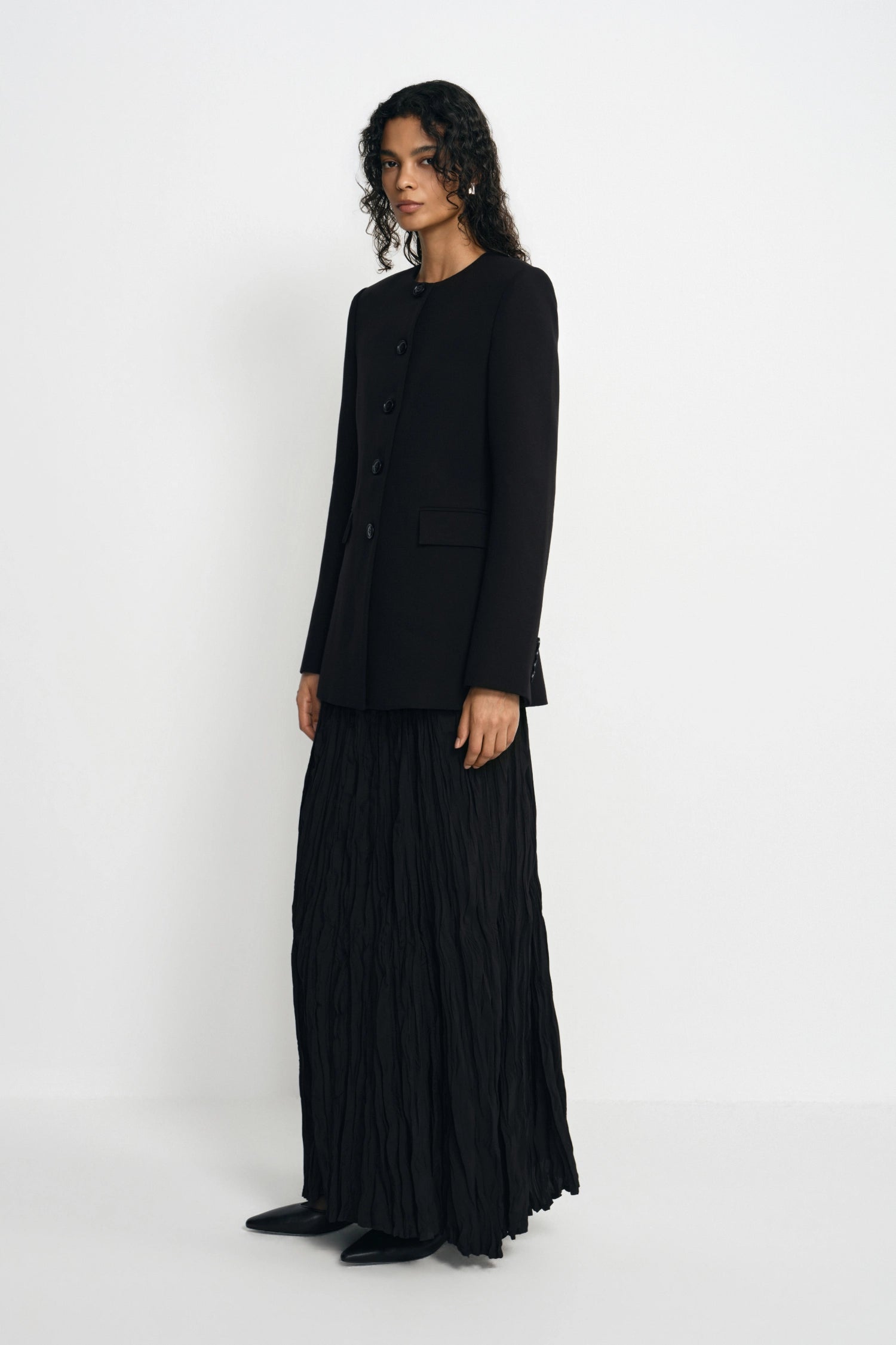 A model stands in a studio wearing a black long-sleeve cropped jacket and a long black pleated skirt, photographed against a plain light gray background.