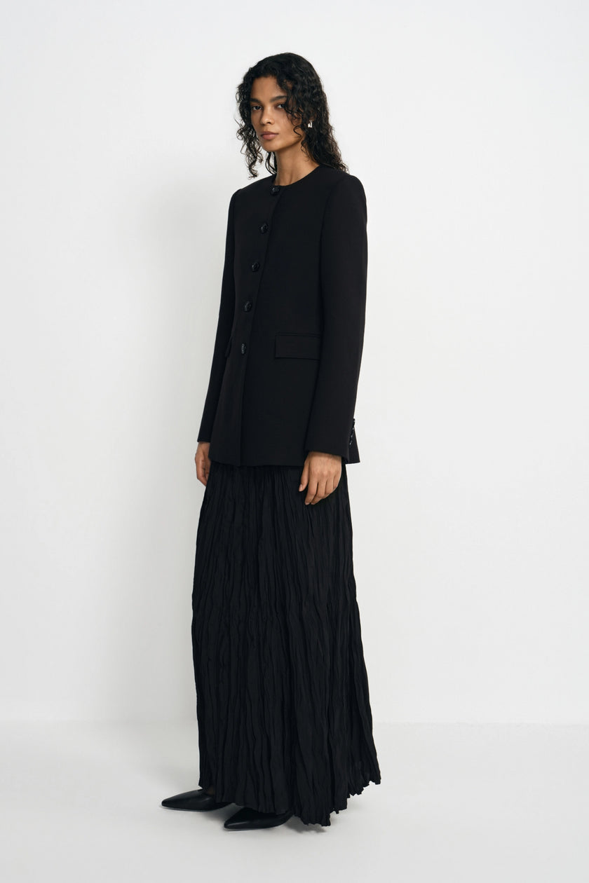 A model stands in a studio wearing a black long-sleeve cropped jacket and a long black pleated skirt, photographed against a plain light gray background.